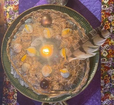 Ecobricolage artwork of a bowl filled with sweet grass, nesting materials, stones, a flame in the centre, and a feather
